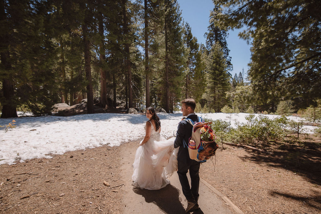 A bride in a wedding dress and a man with a colorful backpack walk along a forest path with snow and trees under a clear blue sky.