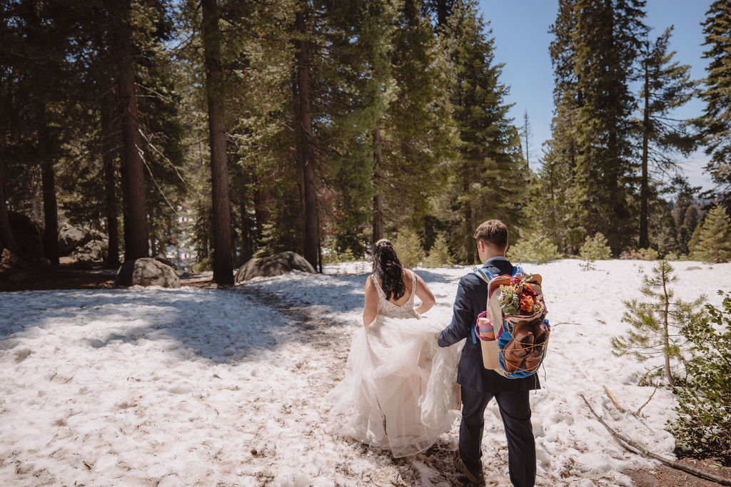 A bride in a wedding dress and a man with a colorful backpack walk along a forest path with snow and trees under a clear blue sky.