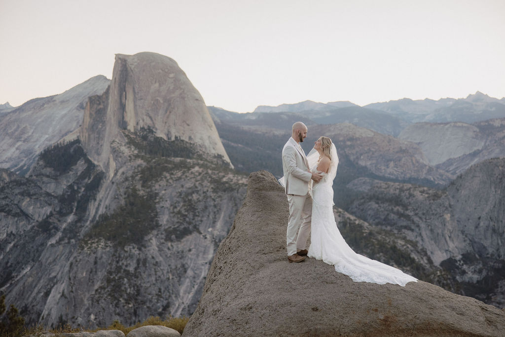 Bride and groom in wedding attire embrace on a mountain overlook with a natural landscape and sunlight in the background.