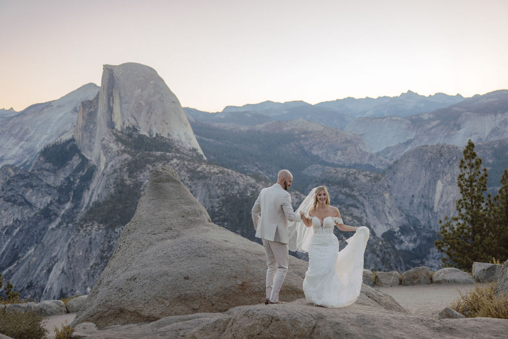 Bride and groom in wedding attire embrace on a mountain overlook with a natural landscape and sunlight in the background.