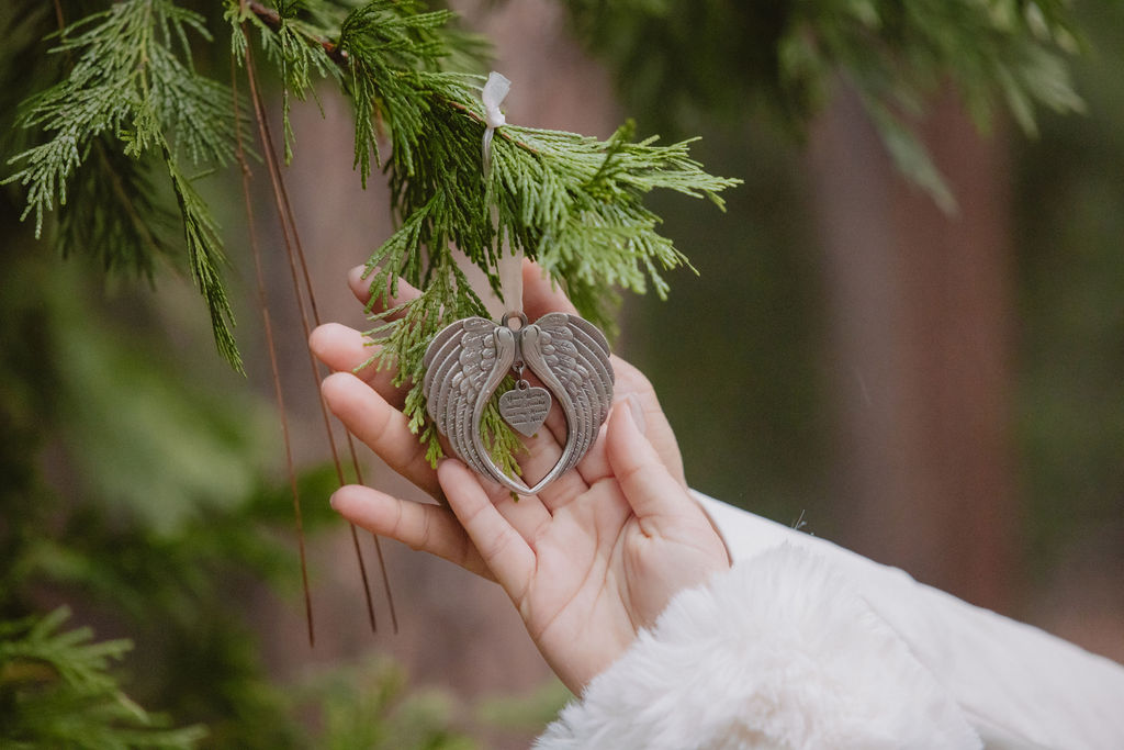 A hand holds a heart-shaped ornament with angel wings against a background of green foliage.