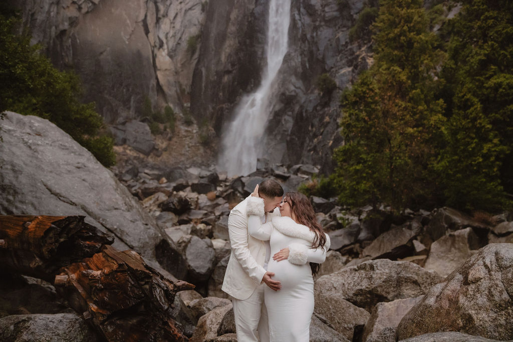 A woman in a white dress holds a baby in front of a rocky landscape with a tall waterfall in the background for a maternity photos in Yosemites