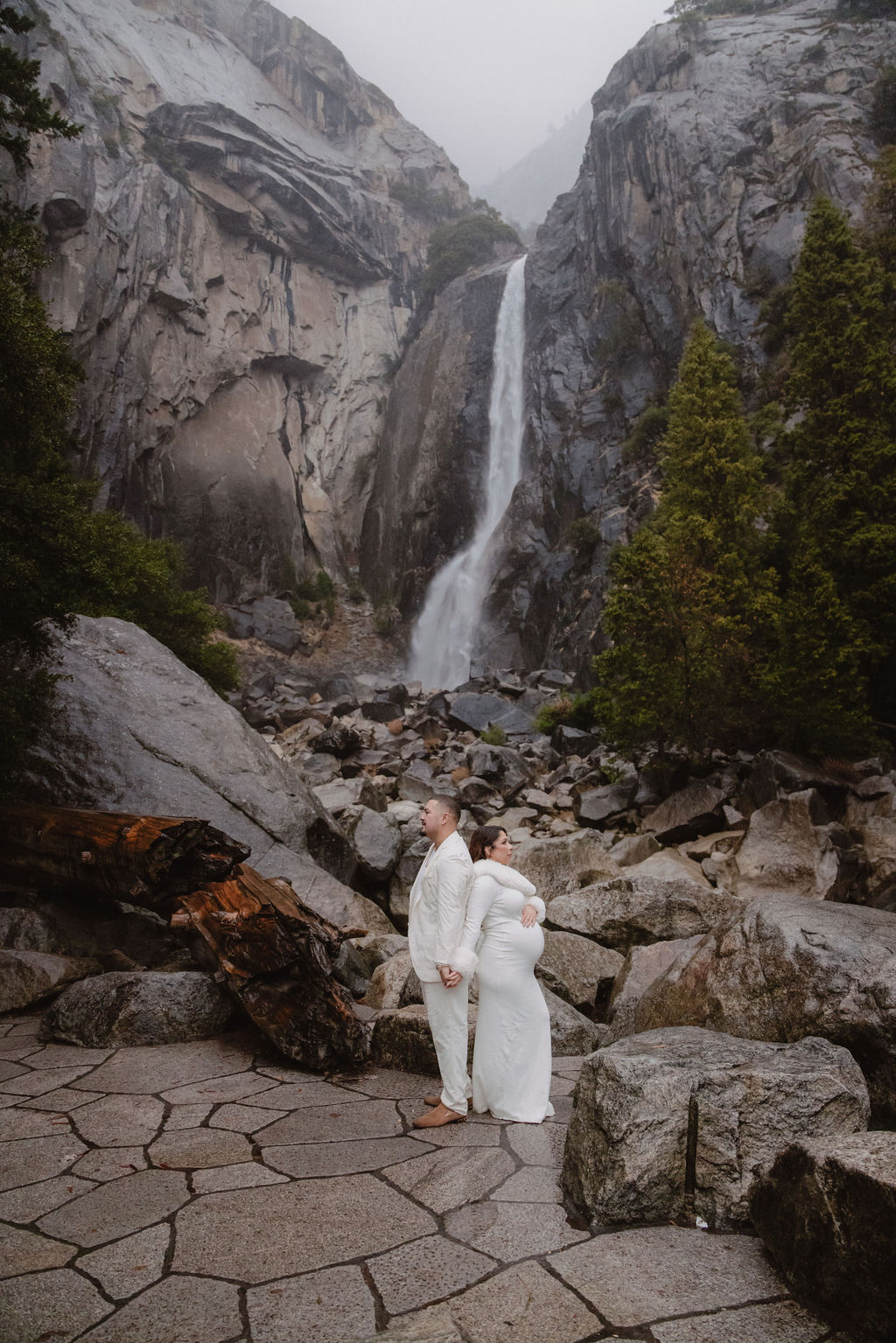 Woman in a white dress stands in front of a rocky landscape with a waterfall in the background.