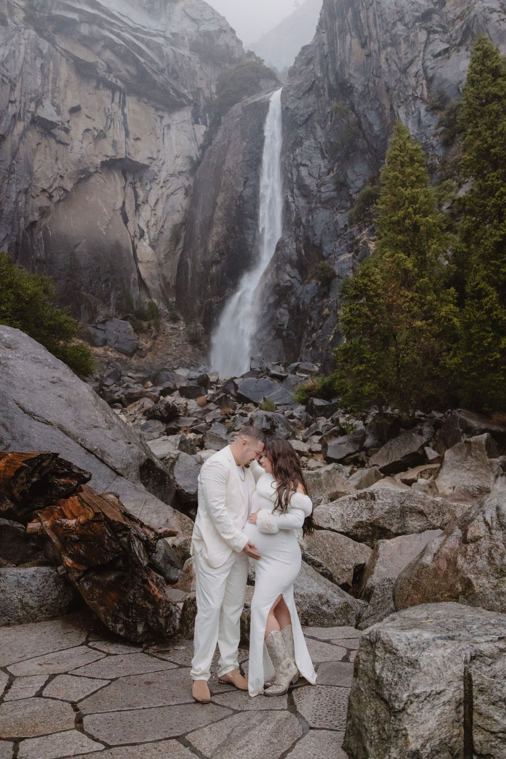 Woman in a white dress stands in front of a rocky landscape with a waterfall in the background.