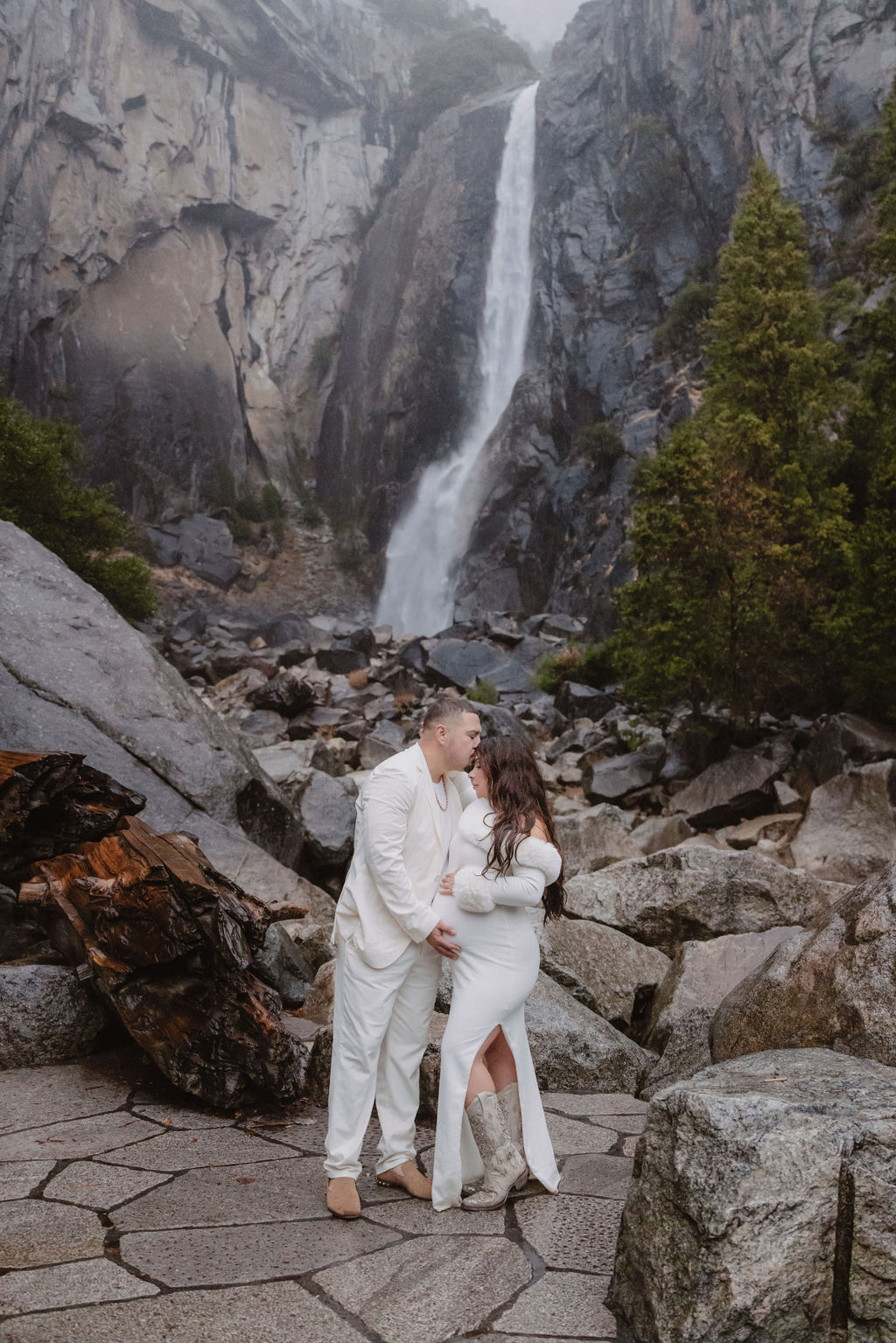 A woman in a white dress holds a baby in front of a rocky landscape with a tall waterfall in the background for a maternity photos in Yosemites