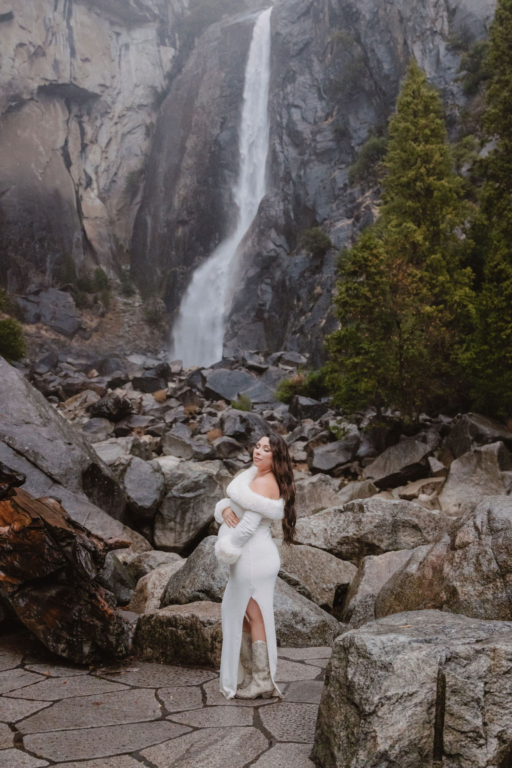 Woman in a white dress stands in front of a rocky landscape with a waterfall in the background.