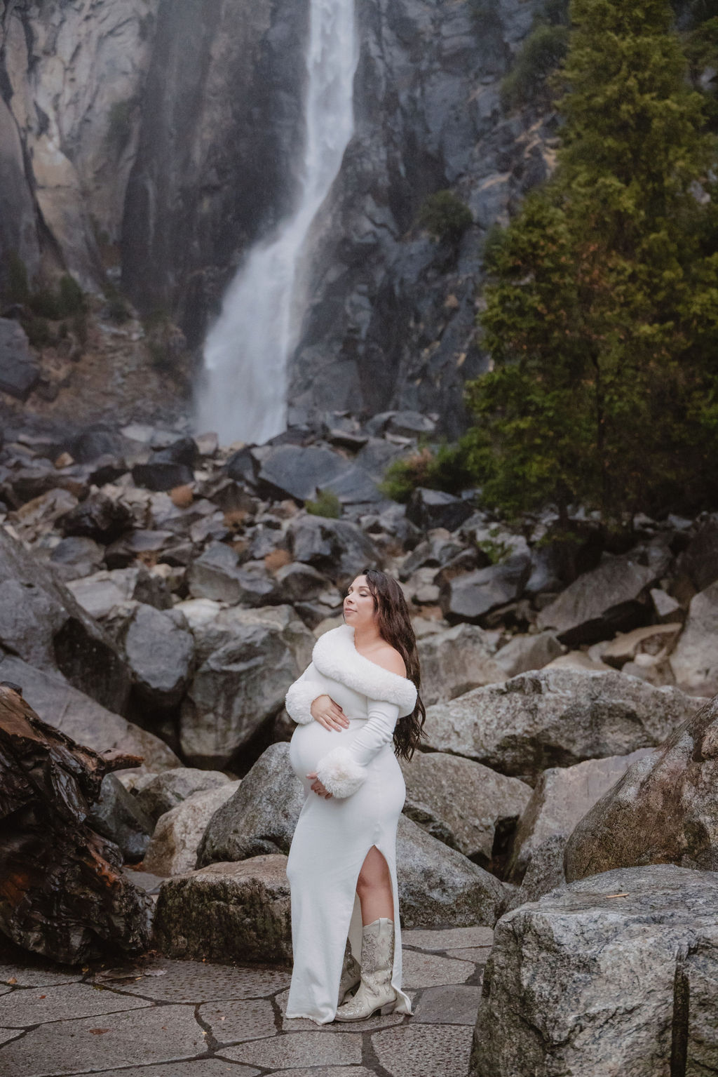 Woman in a white dress stands in front of a rocky landscape with a waterfall in the background.