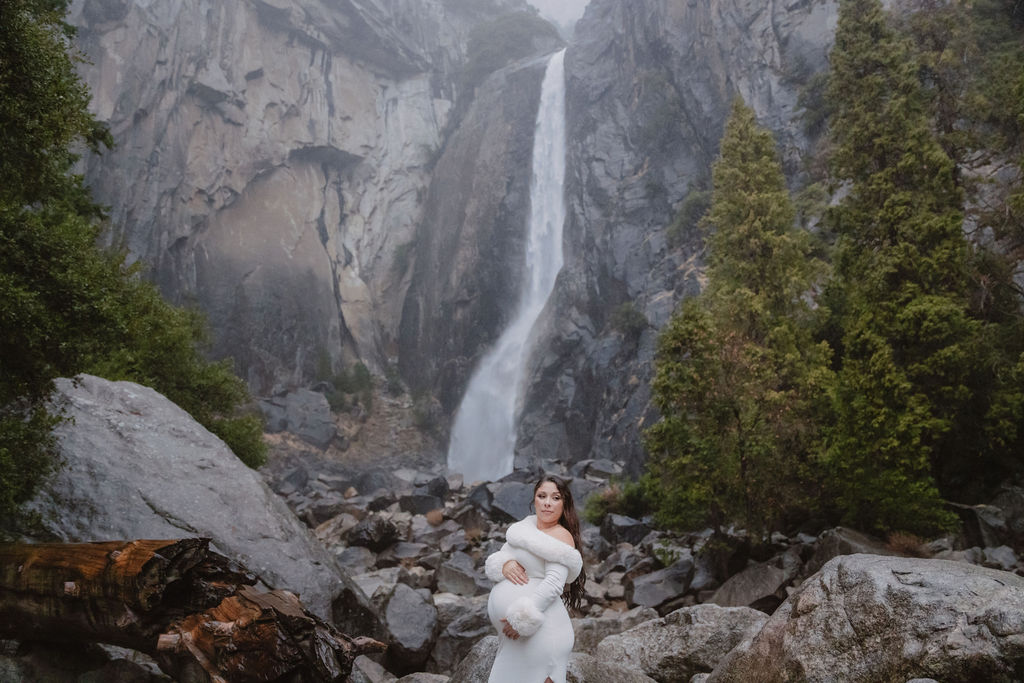 Person in a white dress standing on rocky terrain with a large waterfall in the background, surrounded by trees and cliffs.