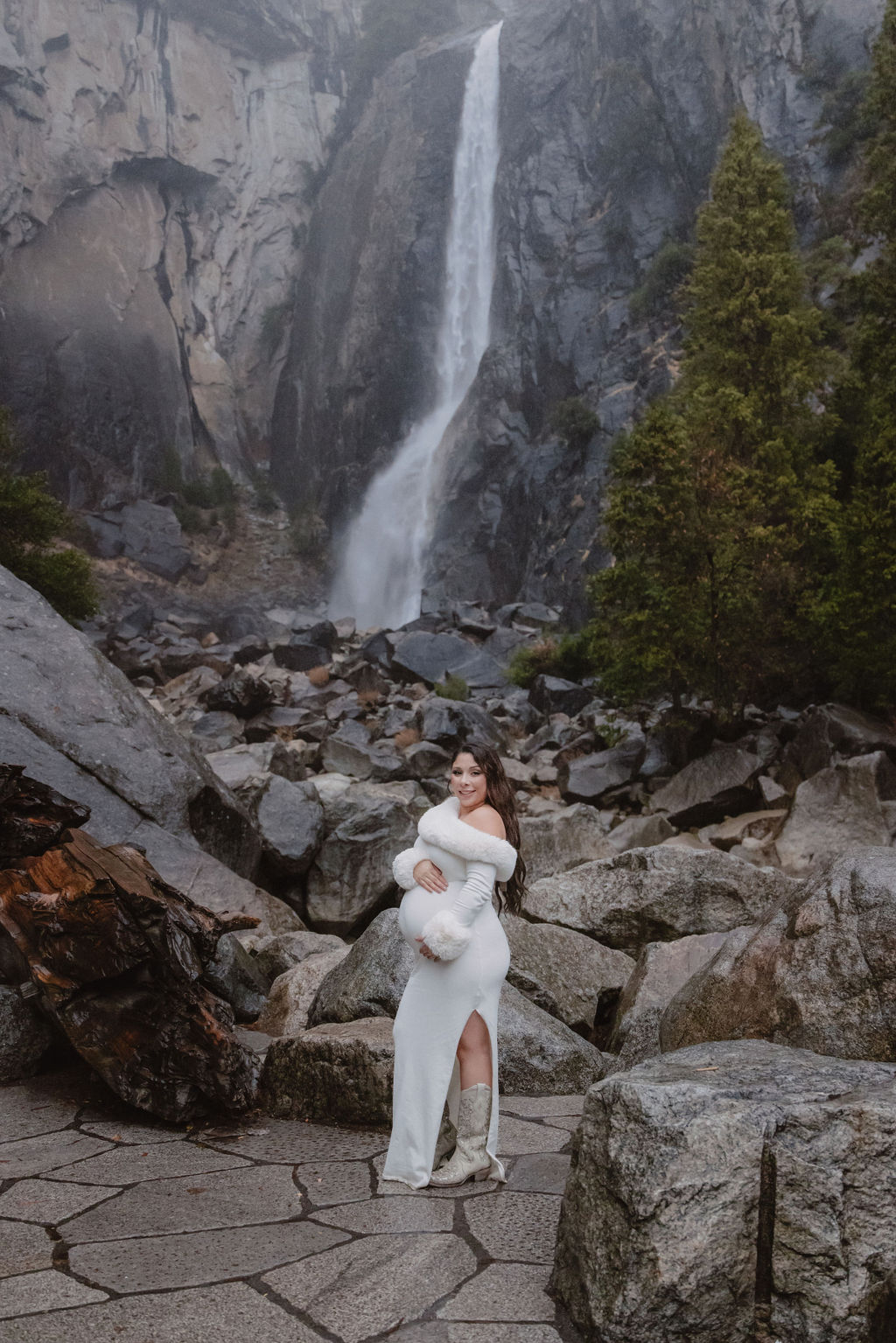 A woman in a white dress holds a baby in front of a rocky landscape with a tall waterfall in the background for a maternity photos in Yosemites