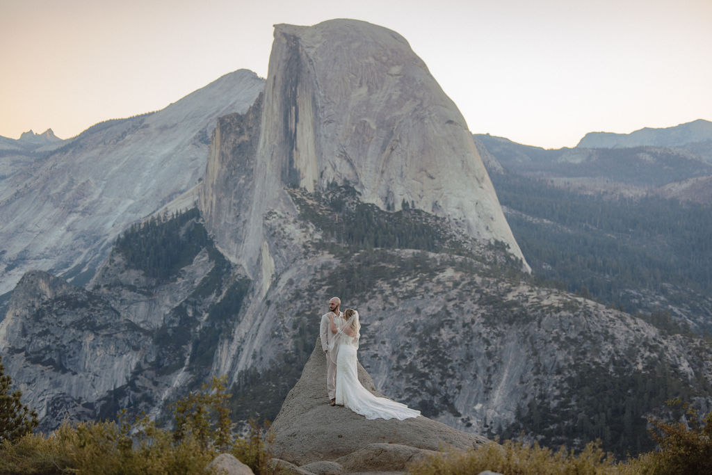 Bride and groom in wedding attire embrace on a mountain overlook with a natural landscape and sunlight in the background.