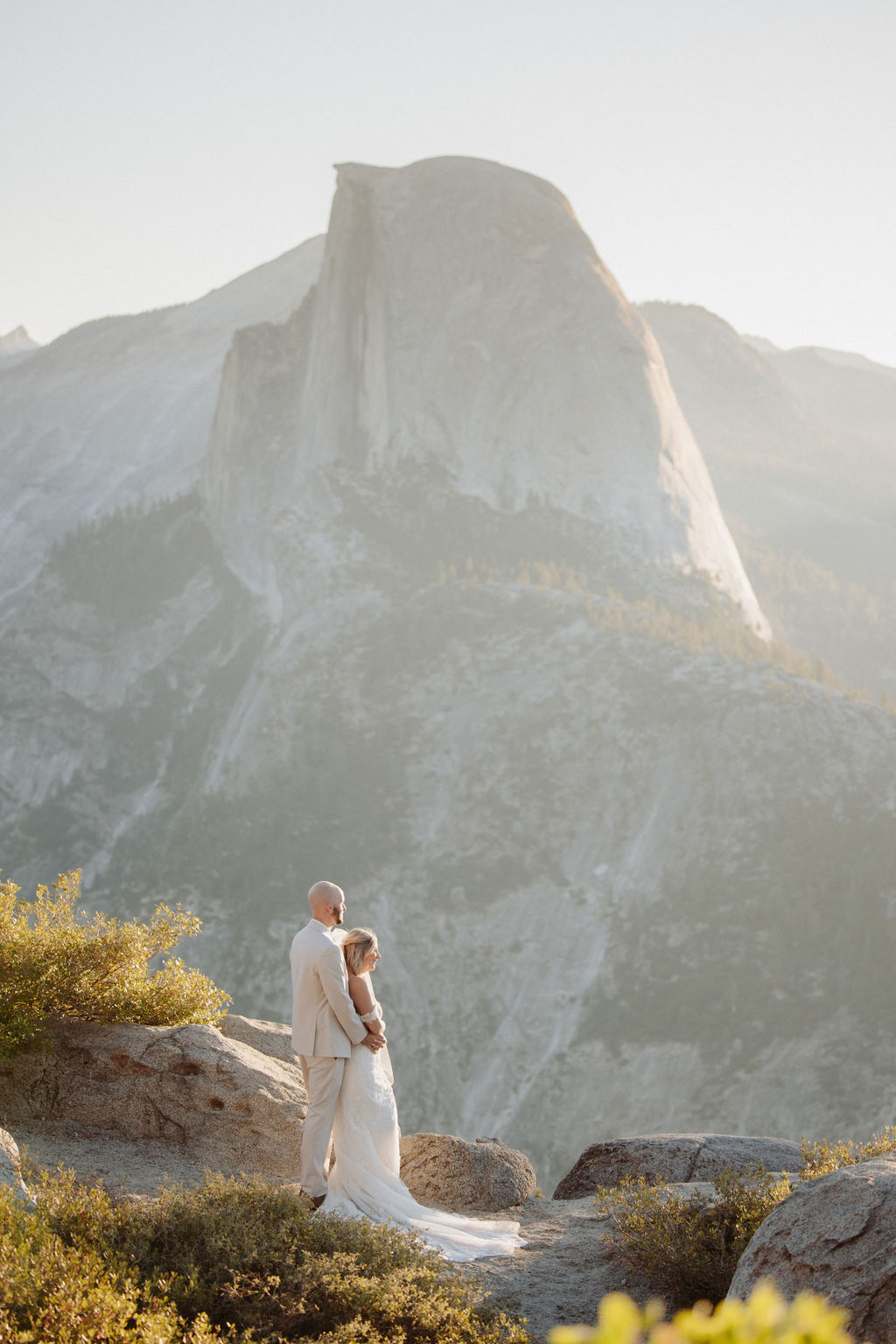 Bride and groom in wedding attire embrace on a mountain overlook with a natural landscape and sunlight in the background.