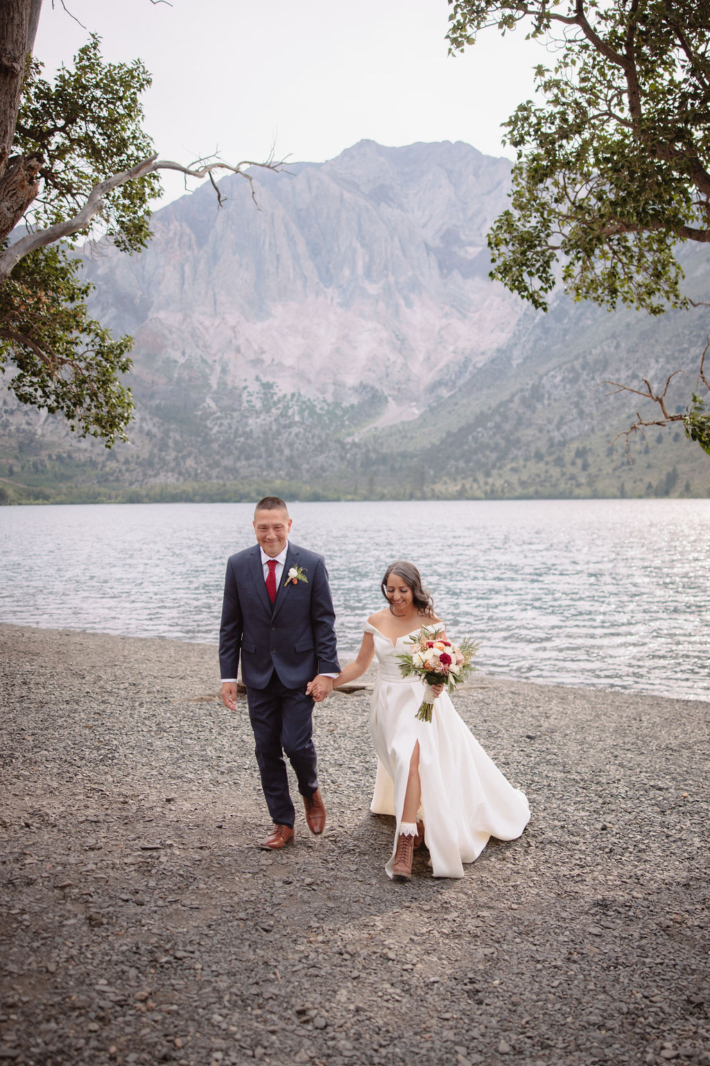 A couple in formal attire walks hand in hand on a rocky lakeside, surrounded by trees and mountains in the background.