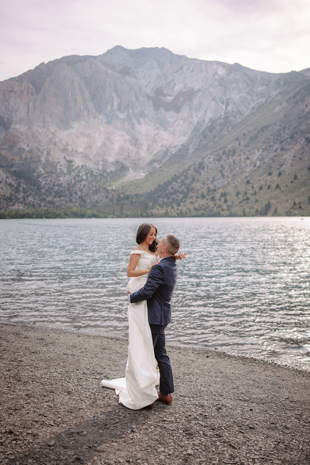 A couple in formal attire walks hand in hand on a rocky lakeside, surrounded by trees and mountains in the background.