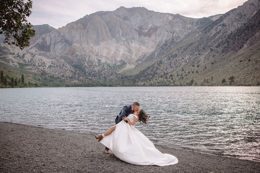 A couple in formal attire walks hand in hand on a rocky lakeside, surrounded by trees and mountains in the background.