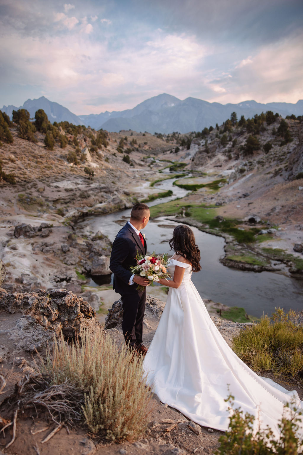 Bride and groom standing on a rocky hillside, looking at a scenic valley with a winding river and distant mountains.