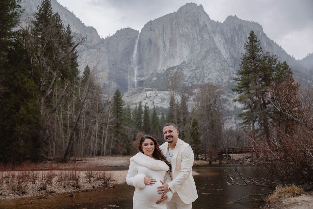 A couple stands together by a river in a forested area, with a large rocky mountain in the background. The woman is in a white dress; the man is in a light-colored suit.