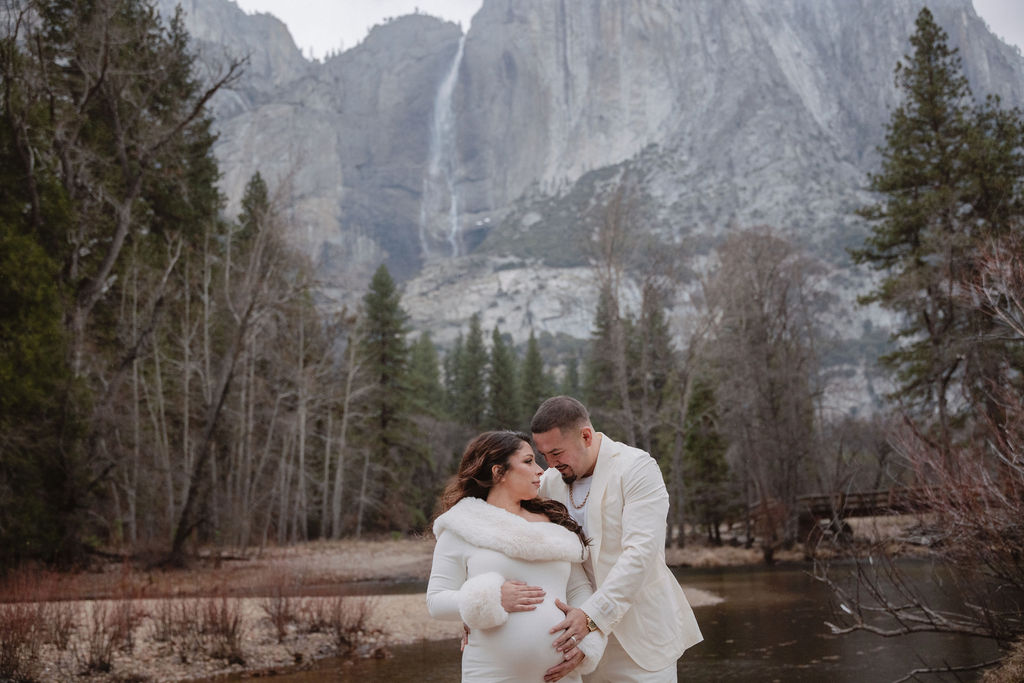 A couple stands together by a river in a forested area, with a large rocky mountain in the background. The woman is in a white dress; the man is in a light-colored suit.