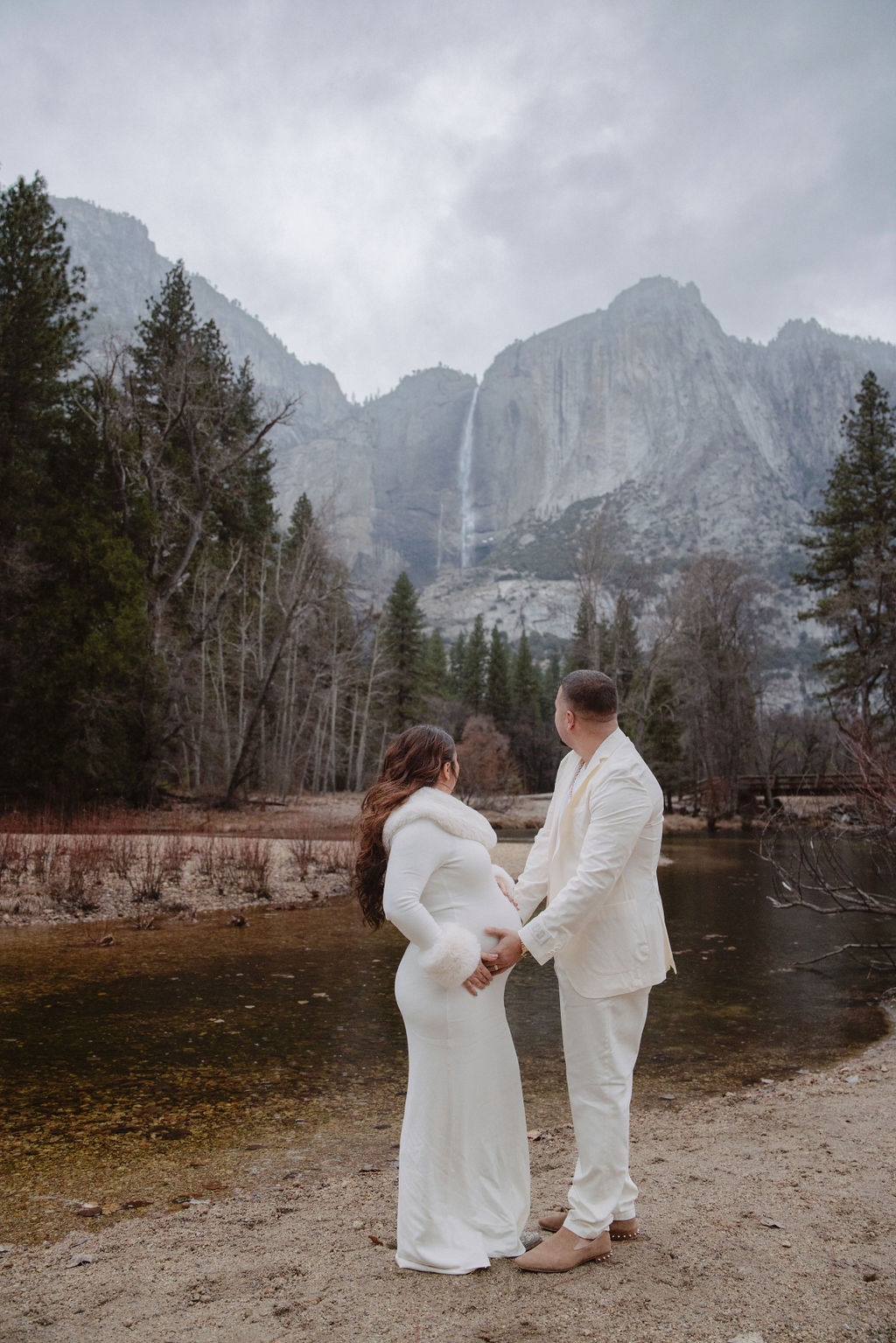 A couple in white attire stands hand in hand by a river, facing a mountain landscape with trees and a waterfall in the background.