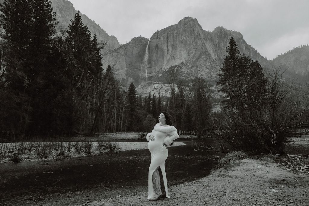 Person in a long dress standing in a natural landscape with trees and mountains in the background, under a cloudy sky.