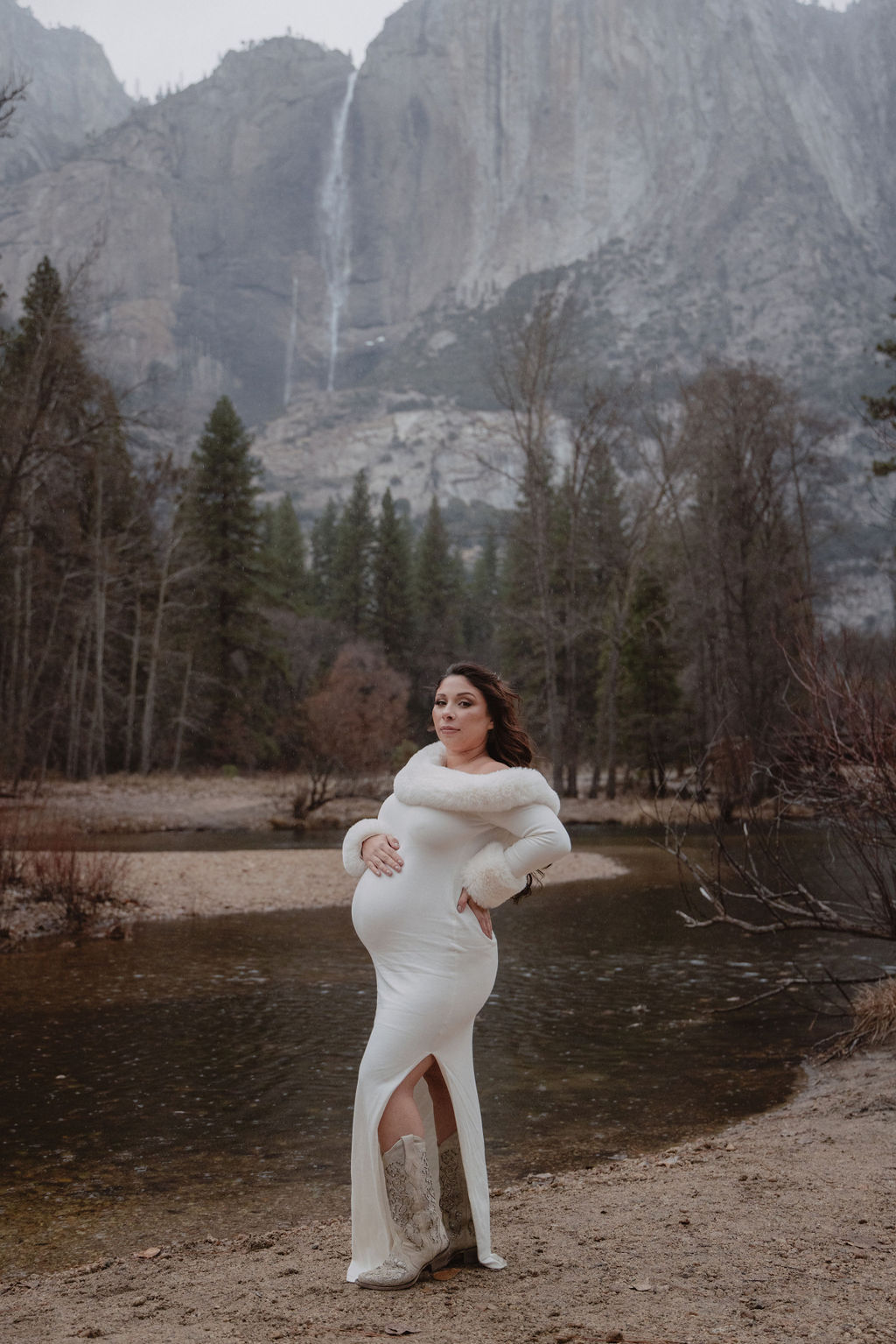 A couple in white clothing walks hand in hand through a grassy field with tall cliffs and cloudy skies in the background for a winter maternity photos in yosemite