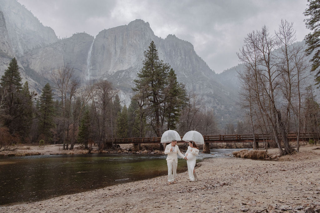 A couple in white clothing walks hand in hand through a grassy field with tall cliffs and cloudy skies in the background for a winter maternity photos in yosemite