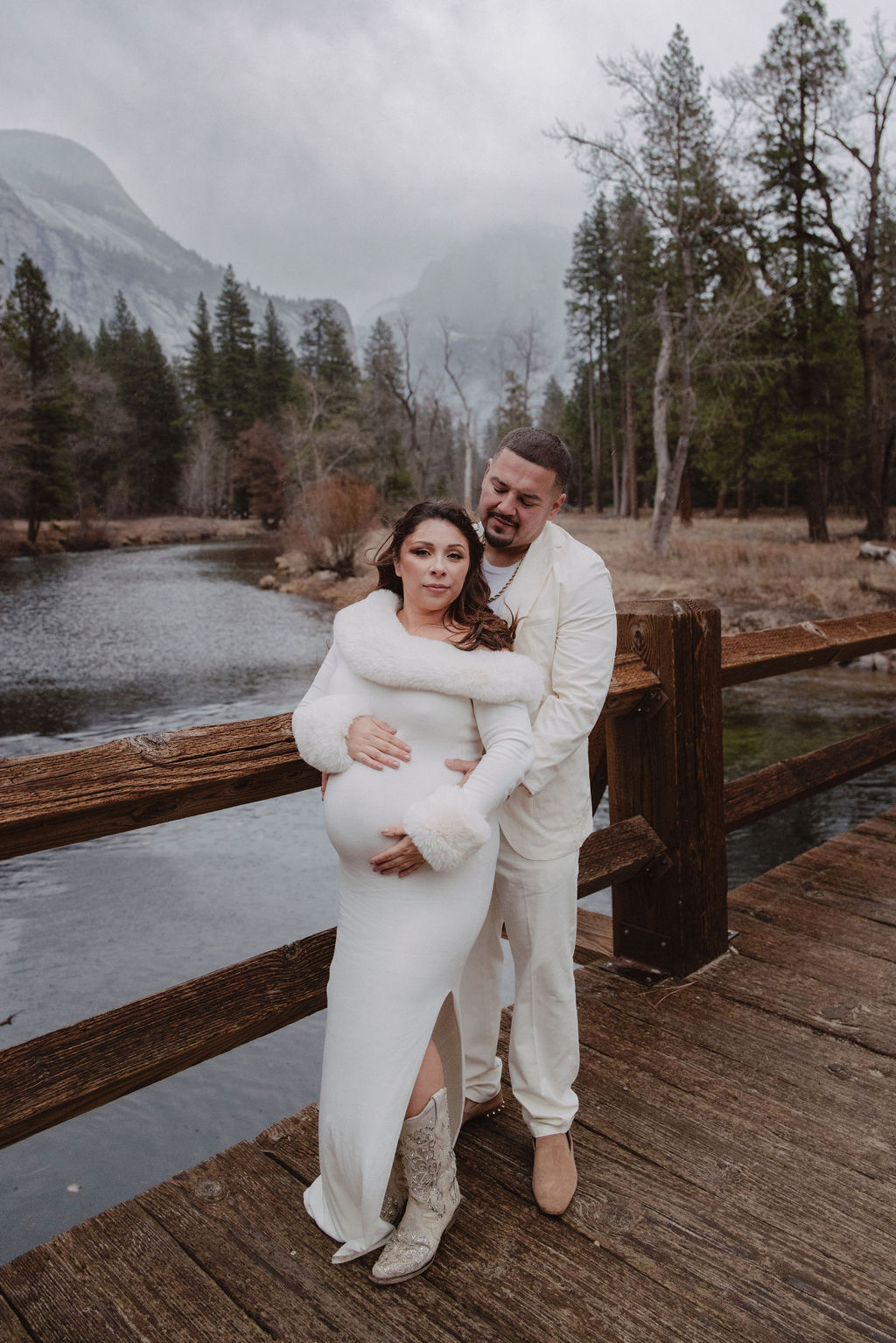 A couple in white clothing walks hand in hand through a grassy field with tall cliffs and cloudy skies in the background for a winter maternity photos in yosemite