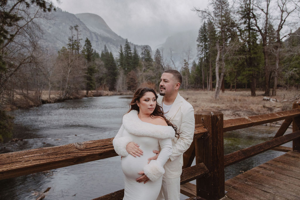 A couple in white clothing walks hand in hand through a grassy field with tall cliffs and cloudy skies in the background for a winter maternity photos in yosemite