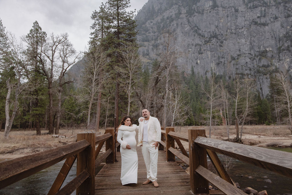 A couple in white clothing walks hand in hand through a grassy field with tall cliffs and cloudy skies in the background for a winter maternity photos in yosemite