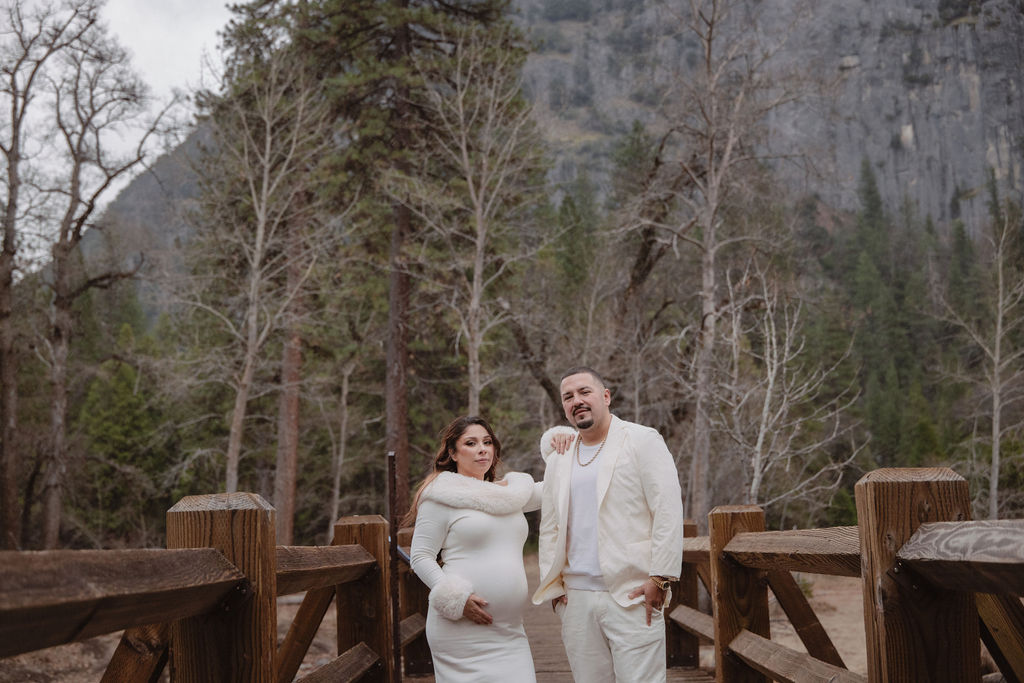 A couple in white clothing walks hand in hand through a grassy field with tall cliffs and cloudy skies in the background for a winter maternity photos in yosemite