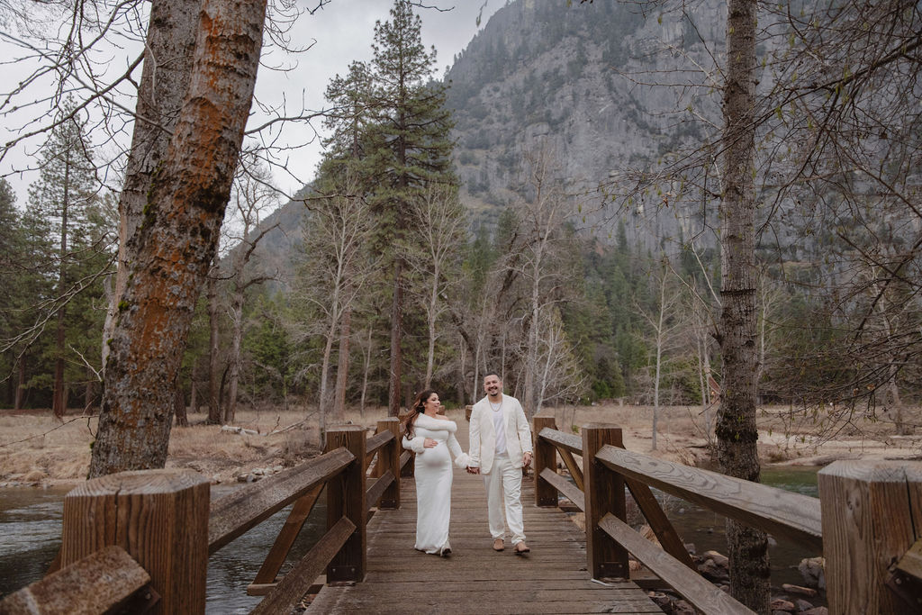 A couple in white clothing walks hand in hand through a grassy field with tall cliffs and cloudy skies in the background for a winter maternity photos in yosemite