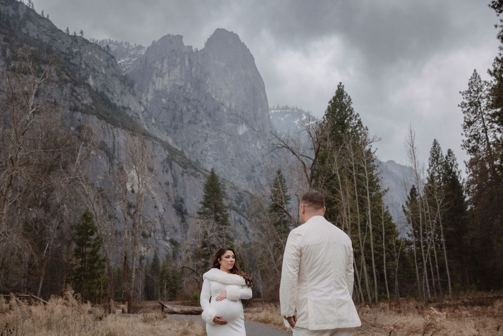 A couple in white clothing walks hand in hand through a grassy field with tall cliffs and cloudy skies in the background for a winter maternity photos in yosemite