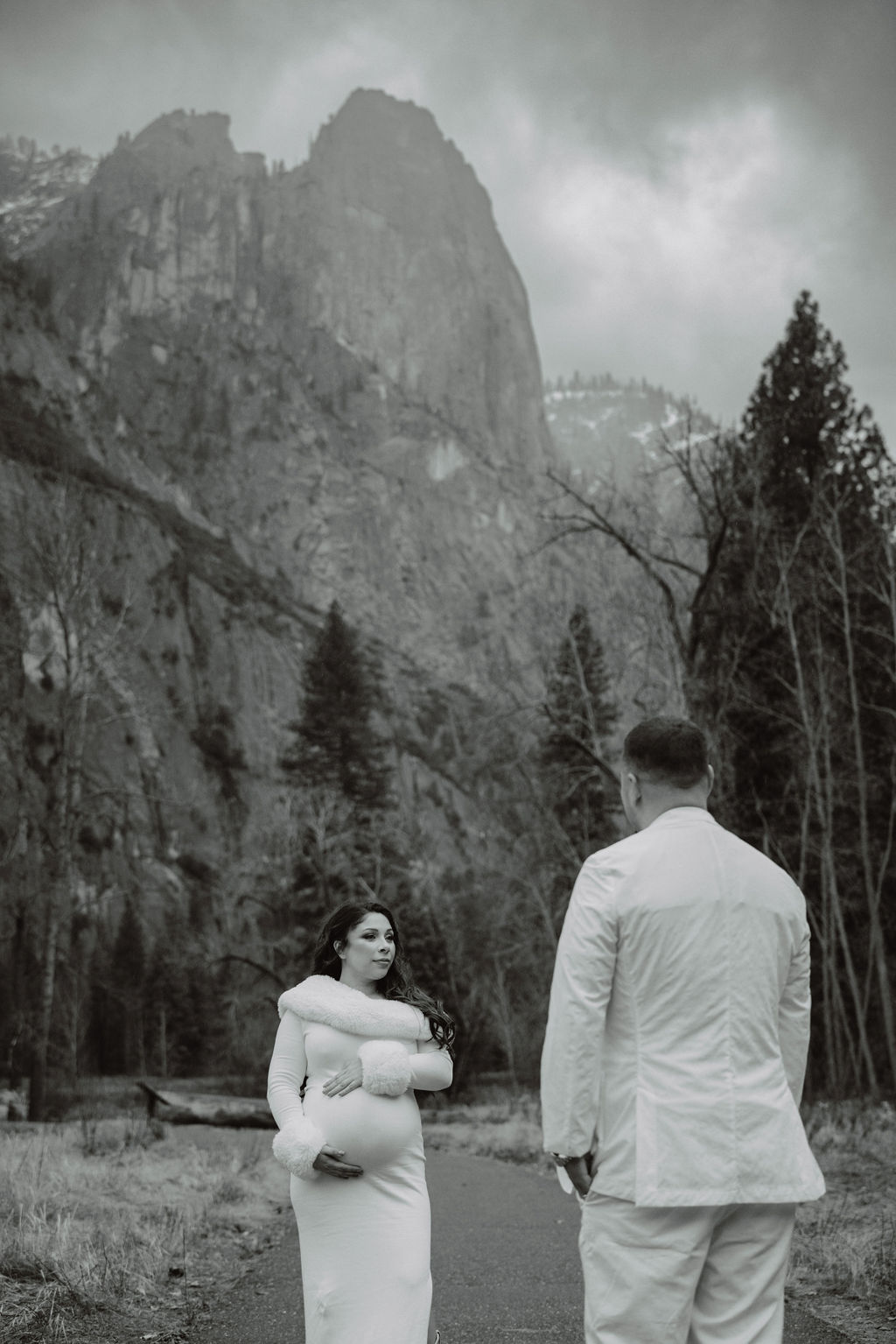 A couple in white clothing walks hand in hand through a grassy field with tall cliffs and cloudy skies in the background for a winter maternity photos in yosemite