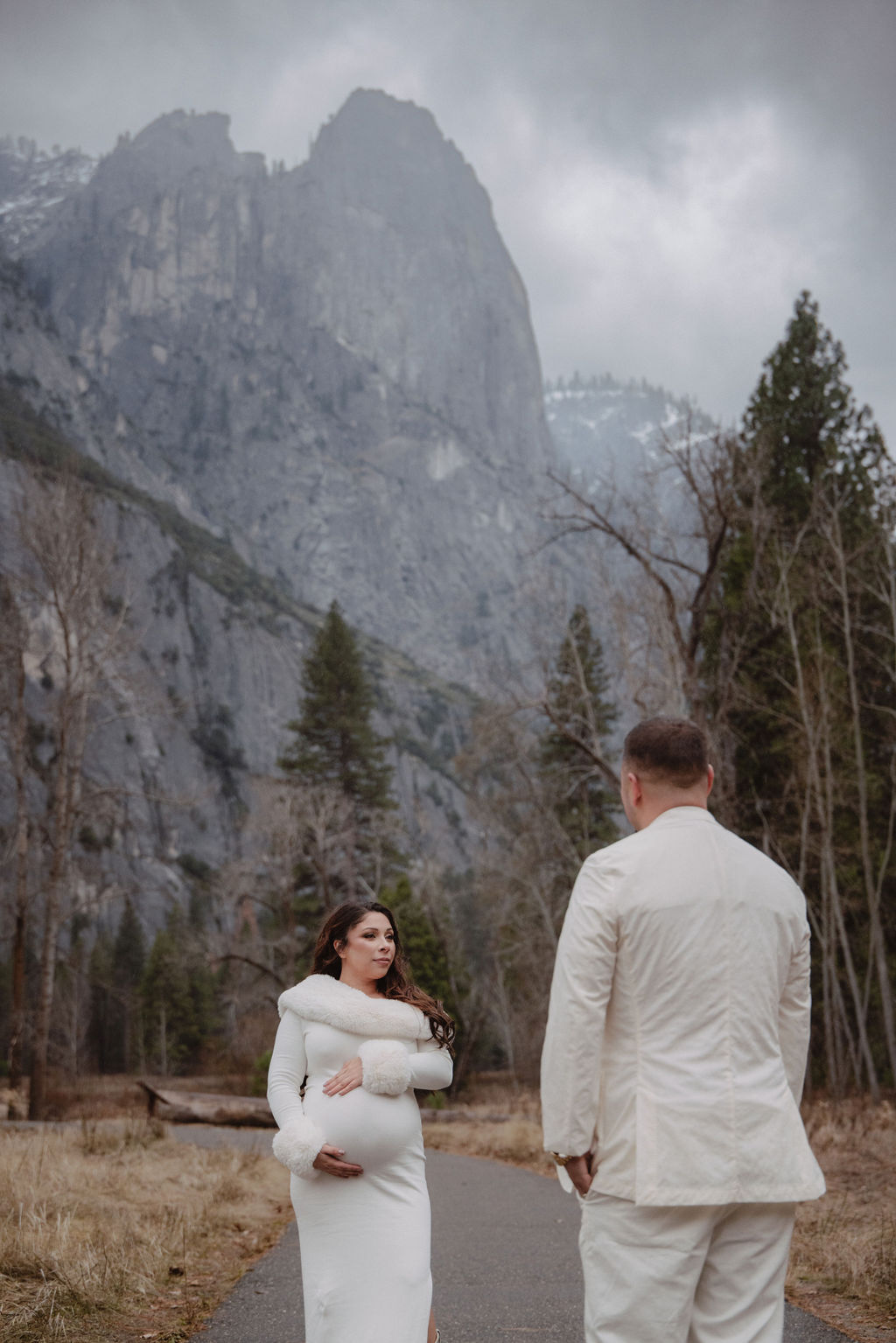 A couple in white clothing walks hand in hand through a grassy field with tall cliffs and cloudy skies in the background for a winter maternity photos in yosemite