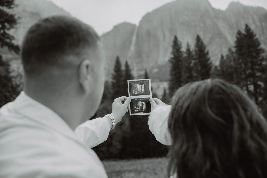 A couple holds two ultrasound photos outdoors, with mountains and trees in the background.