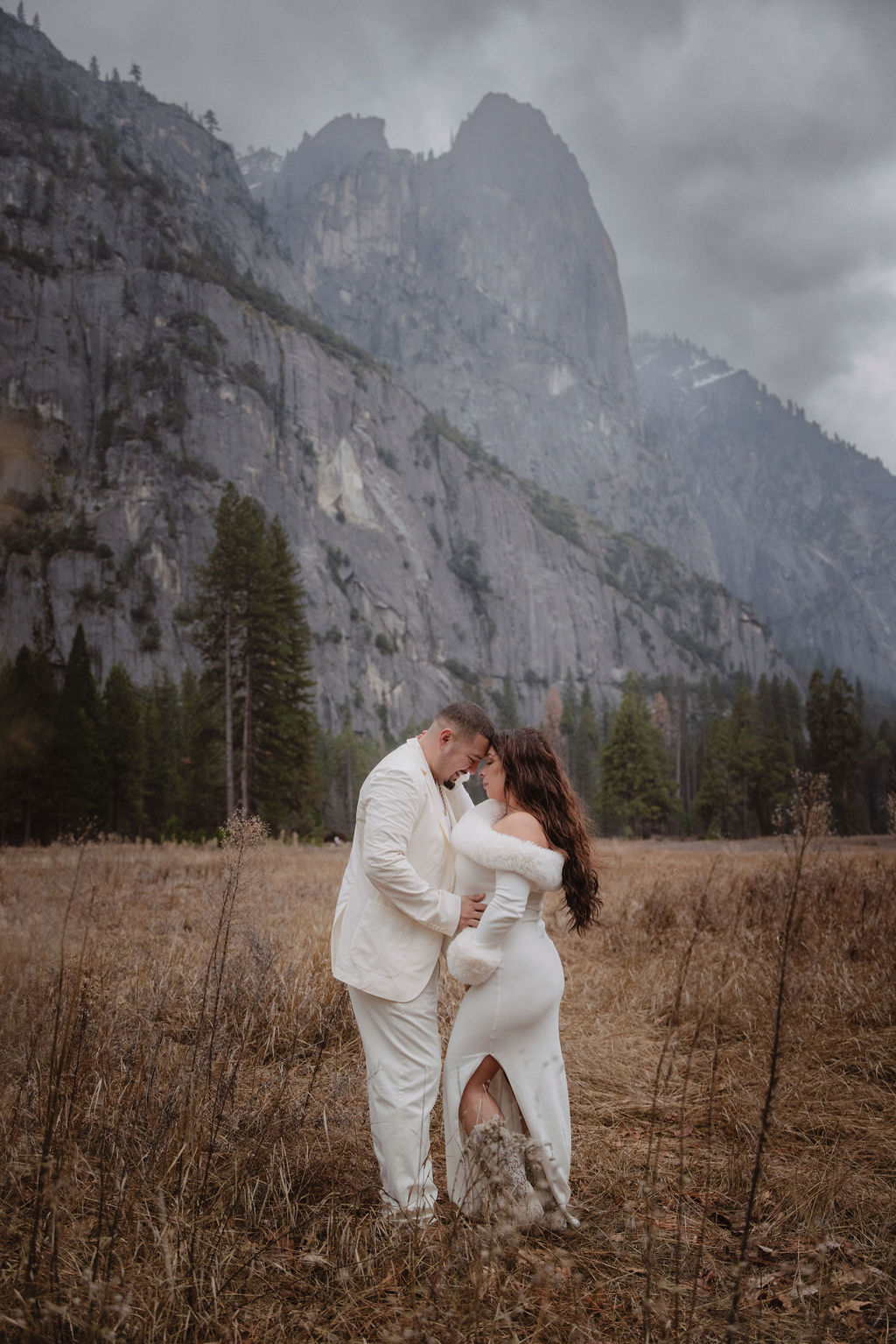 A couple in white clothing walks hand in hand through a grassy field with tall cliffs and cloudy skies in the background for a winter maternity photos in yosemite