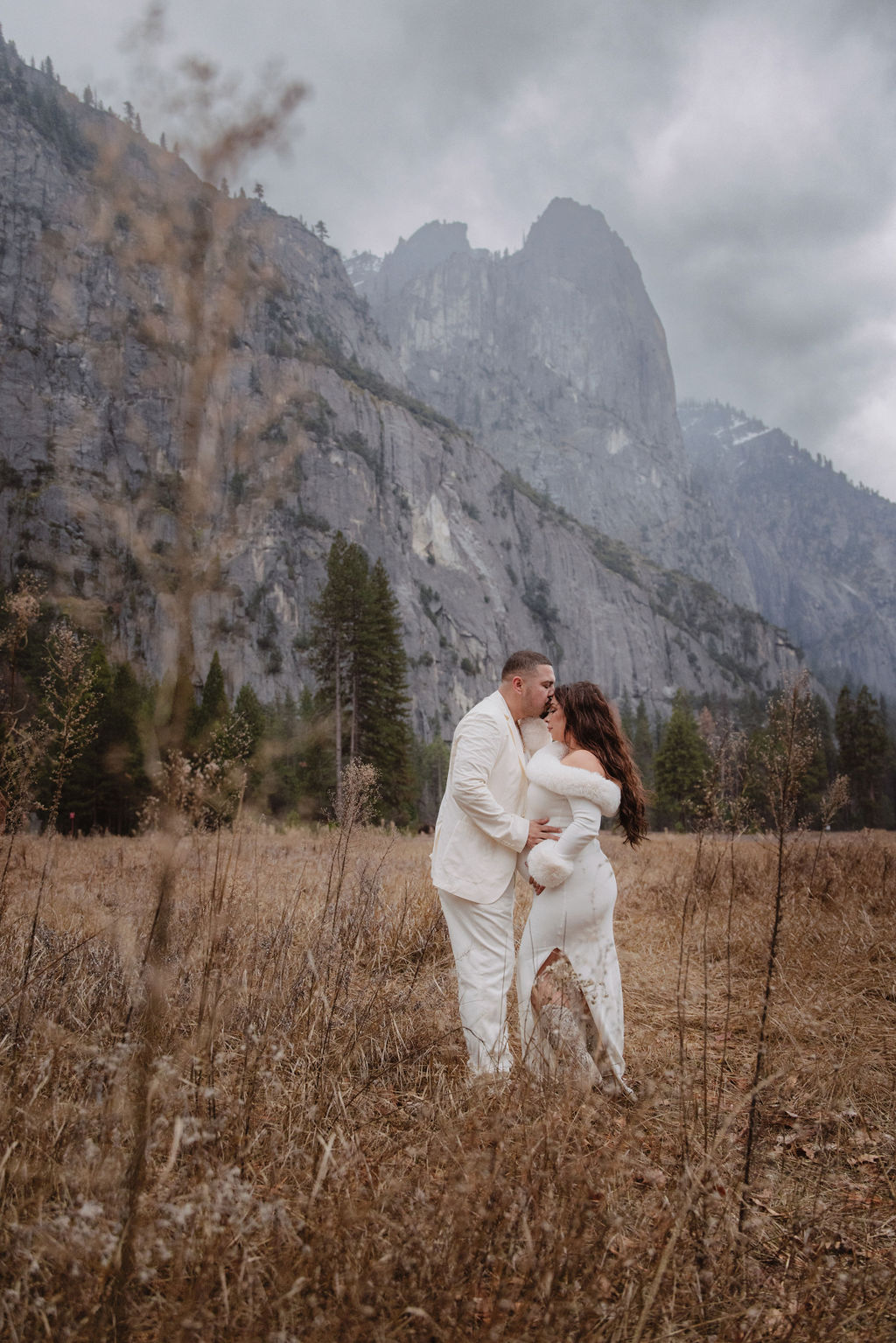 A couple in white clothing walks hand in hand through a grassy field with tall cliffs and cloudy skies in the background for a winter maternity photos in yosemite