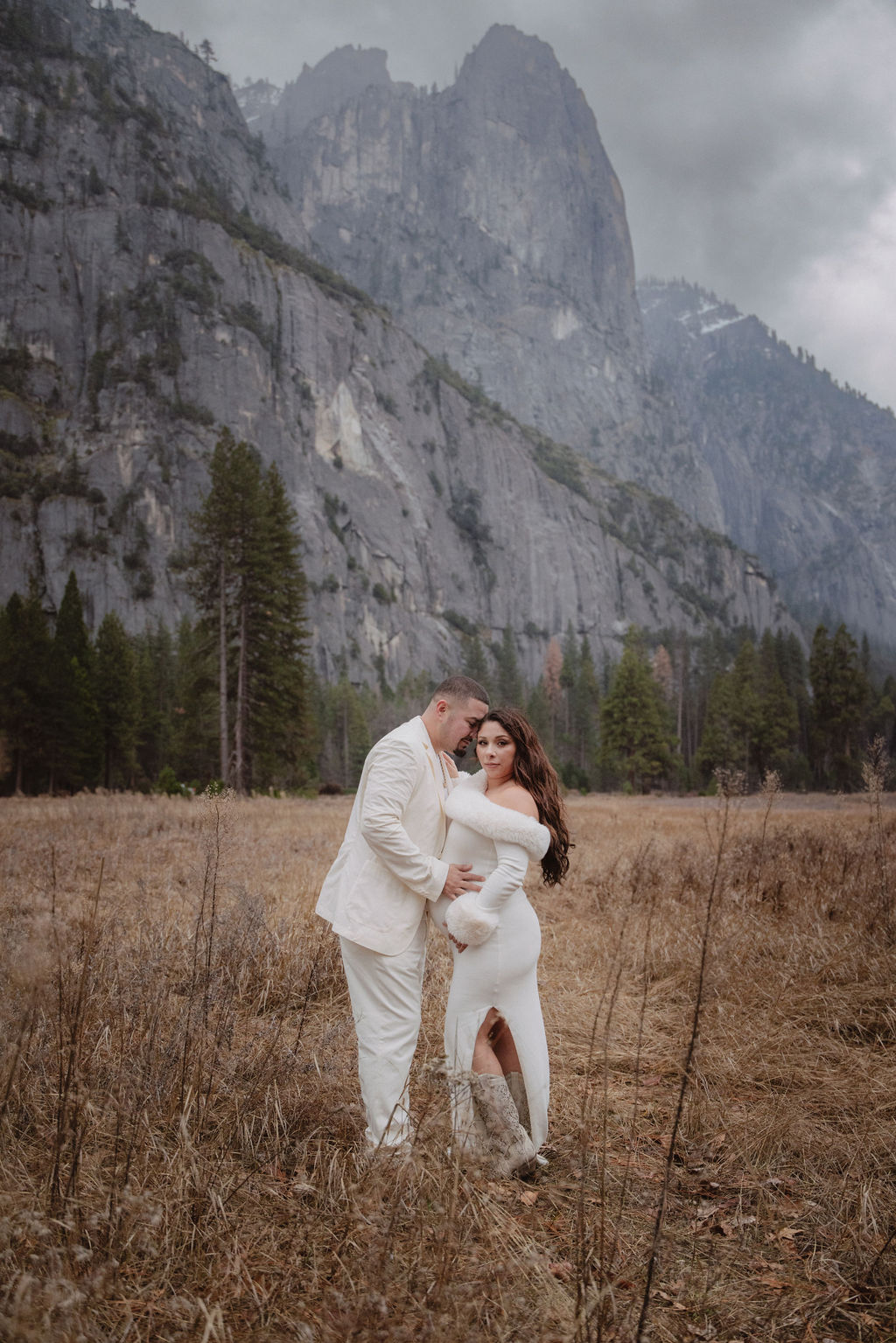 A couple in white clothing walks hand in hand through a grassy field with tall cliffs and cloudy skies in the background for a winter maternity photos in yosemite