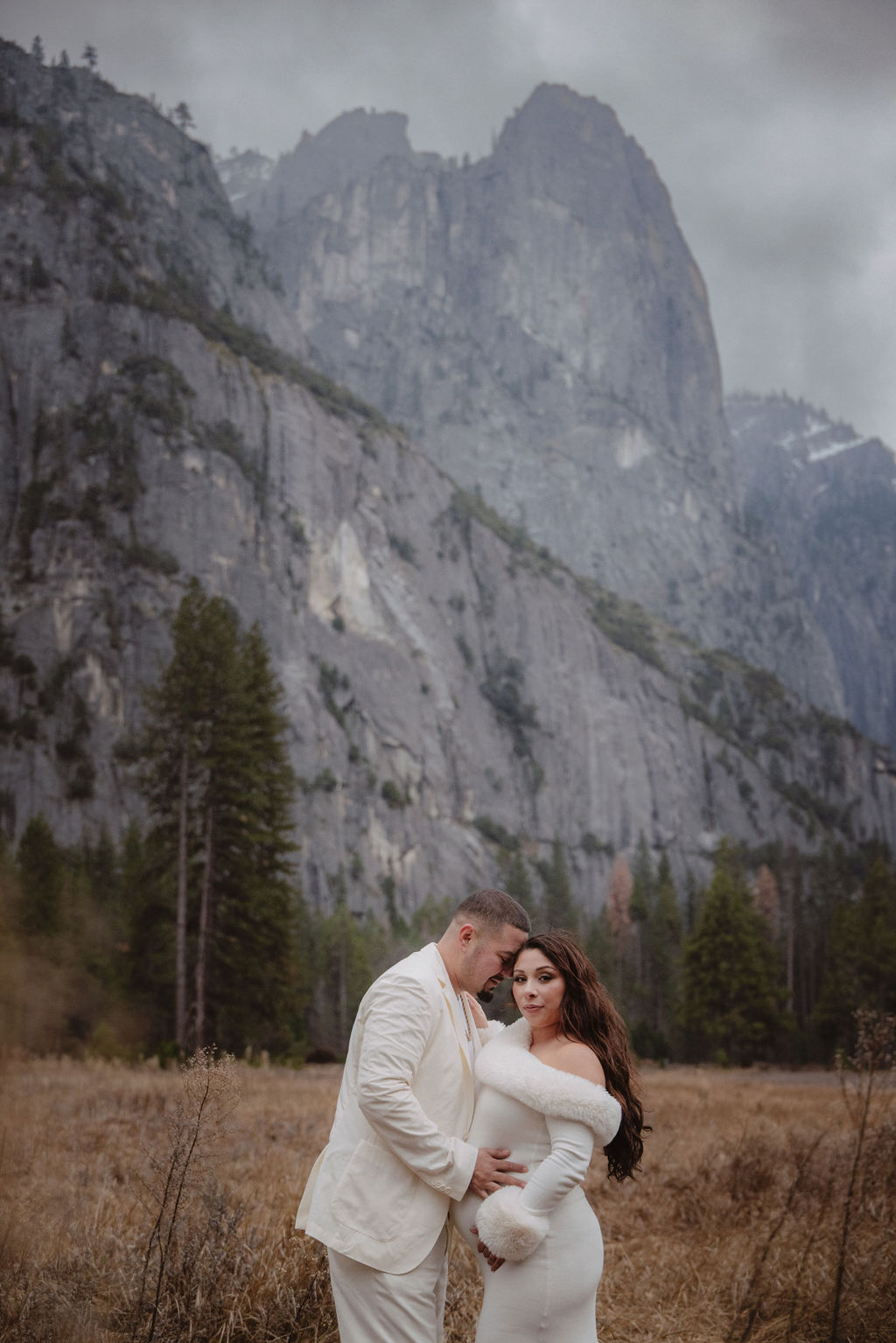 A couple in white clothing walks hand in hand through a grassy field with tall cliffs and cloudy skies in the background for a winter maternity photos in yosemite