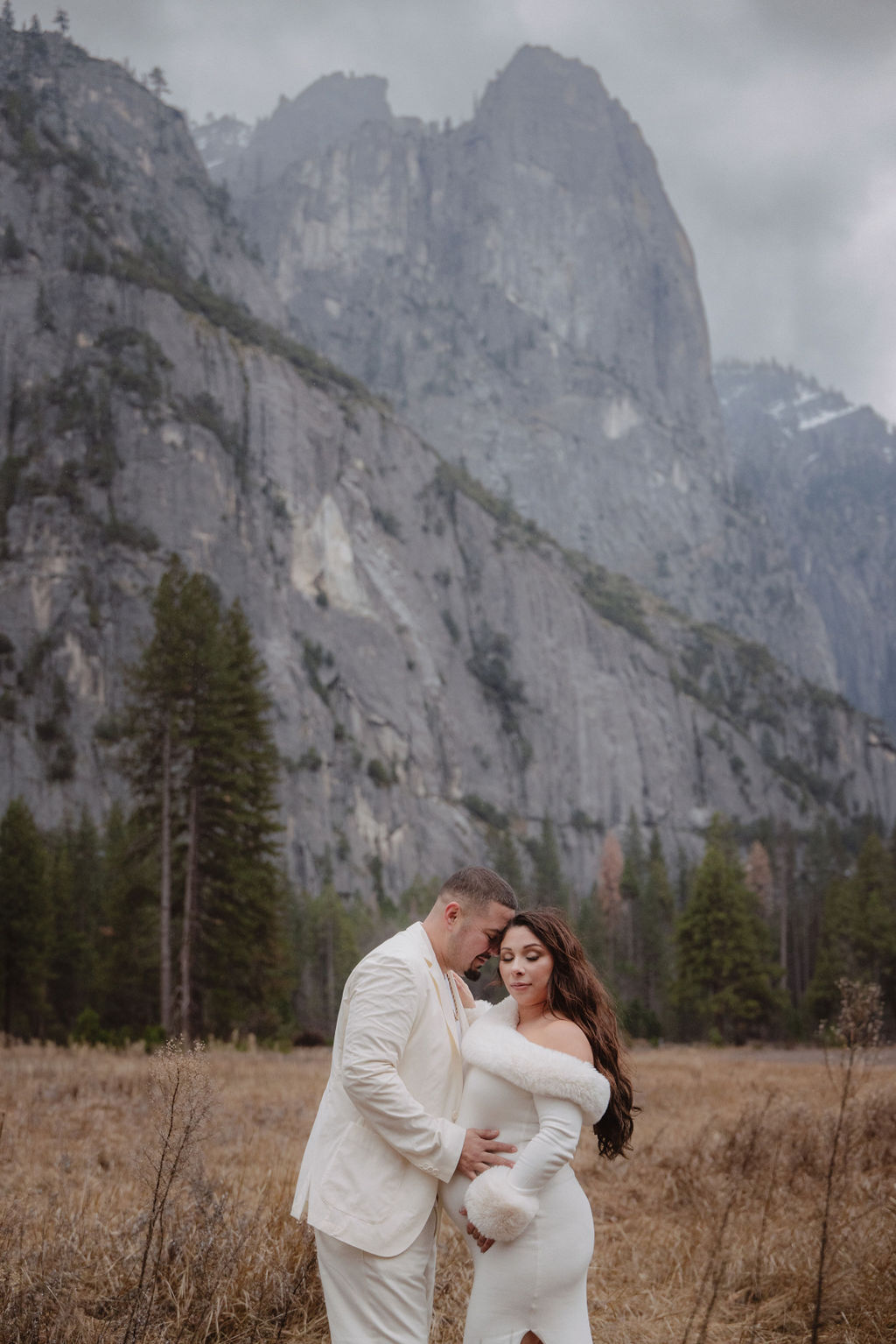 A couple in white clothing walks hand in hand through a grassy field with tall cliffs and cloudy skies in the background for a winter maternity photos in yosemite