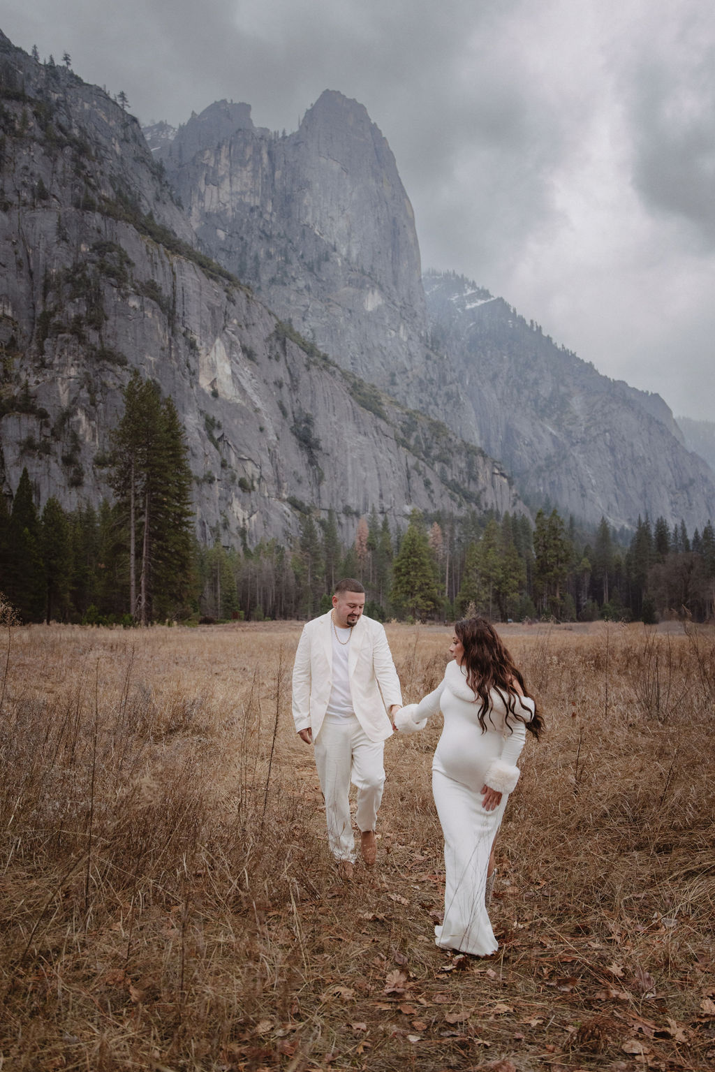 A couple in white clothing walks hand in hand through a grassy field with tall cliffs and cloudy skies in the background for a winter maternity photos in yosemite