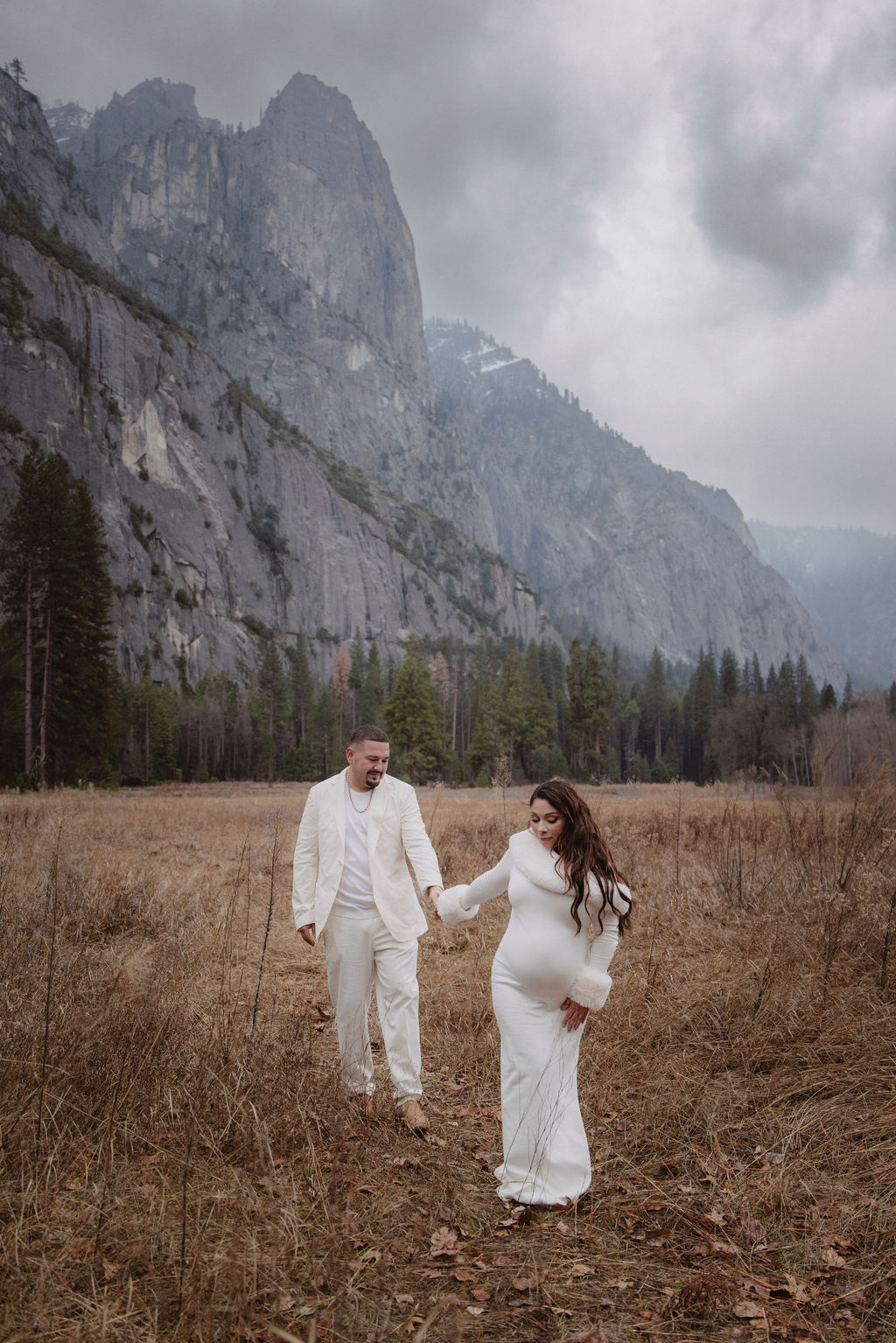 A couple in white clothing walks hand in hand through a grassy field with tall cliffs and cloudy skies in the background for a winter maternity photos in yosemite