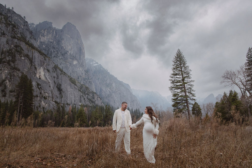 A couple in white clothing walks hand in hand through a grassy field with tall cliffs and cloudy skies in the background for a winter maternity photos in yosemite