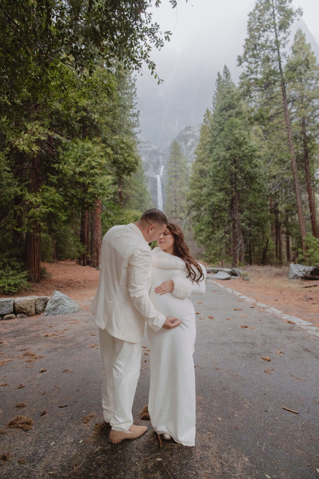 A couple in white attire embrace on a wet, tree-lined path with a waterfall in the background.