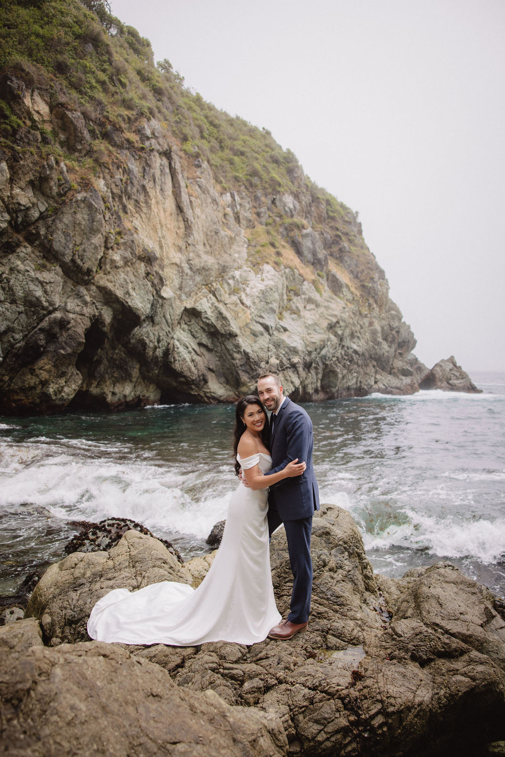 A couple in formal attire poses on rocky seaside terrain with ocean waves and a large cliff in the background for an elopement in California