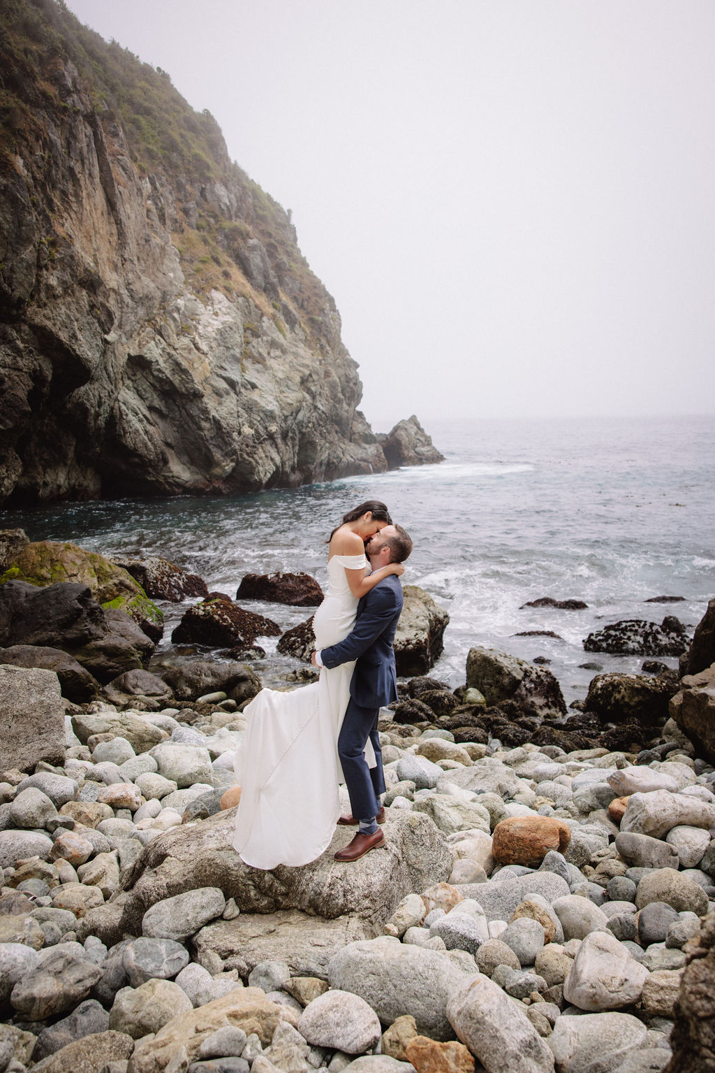 A couple in formal attire poses on rocky seaside terrain with ocean waves and a large cliff in the background for an elopement in California 