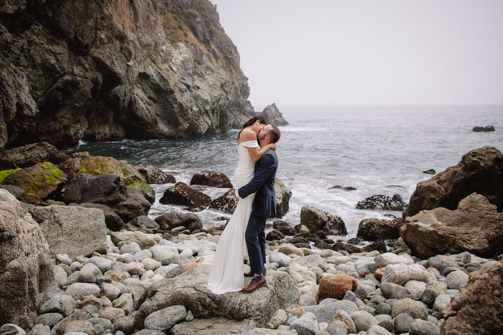 A couple in formal attire poses on rocky seaside terrain with ocean waves and a large cliff in the background for an elopement in California 