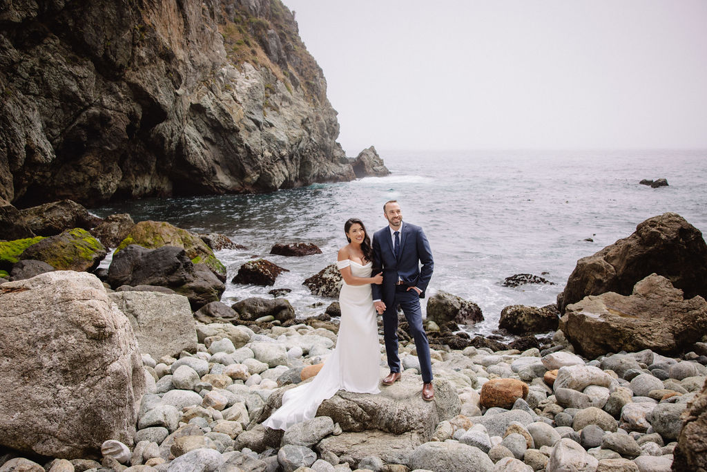 A couple in formal attire poses on rocky seaside terrain with ocean waves and a large cliff in the background for an elopement in California 