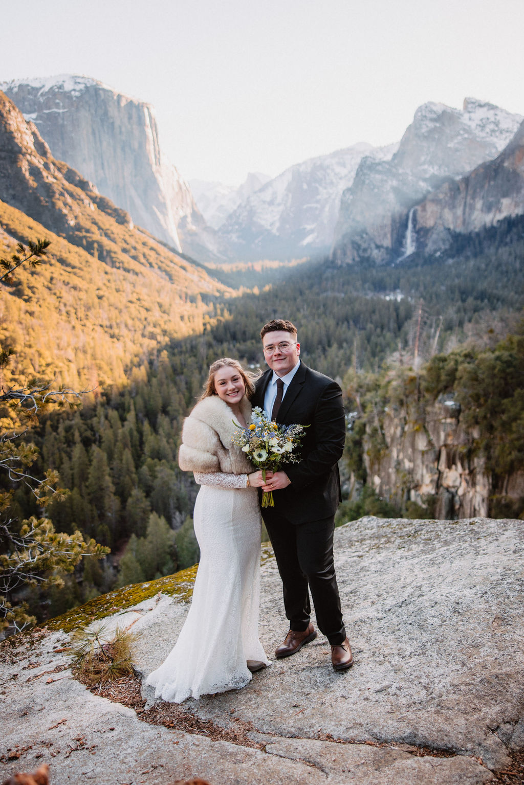 A couple in wedding attire stands smiling on a rock with a mountainous landscape in the background. How to Include Friends & Family in Your Elopement