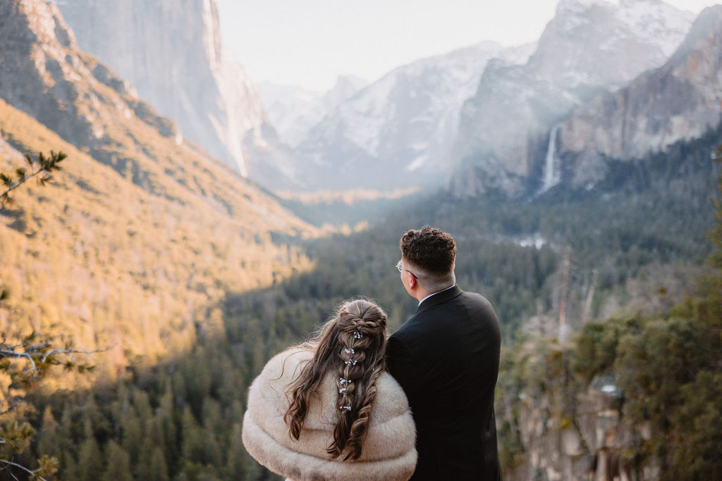 A couple in wedding attire stands smiling on a rock with a mountainous landscape in the background. How to Include Friends & Family in Your Elopement