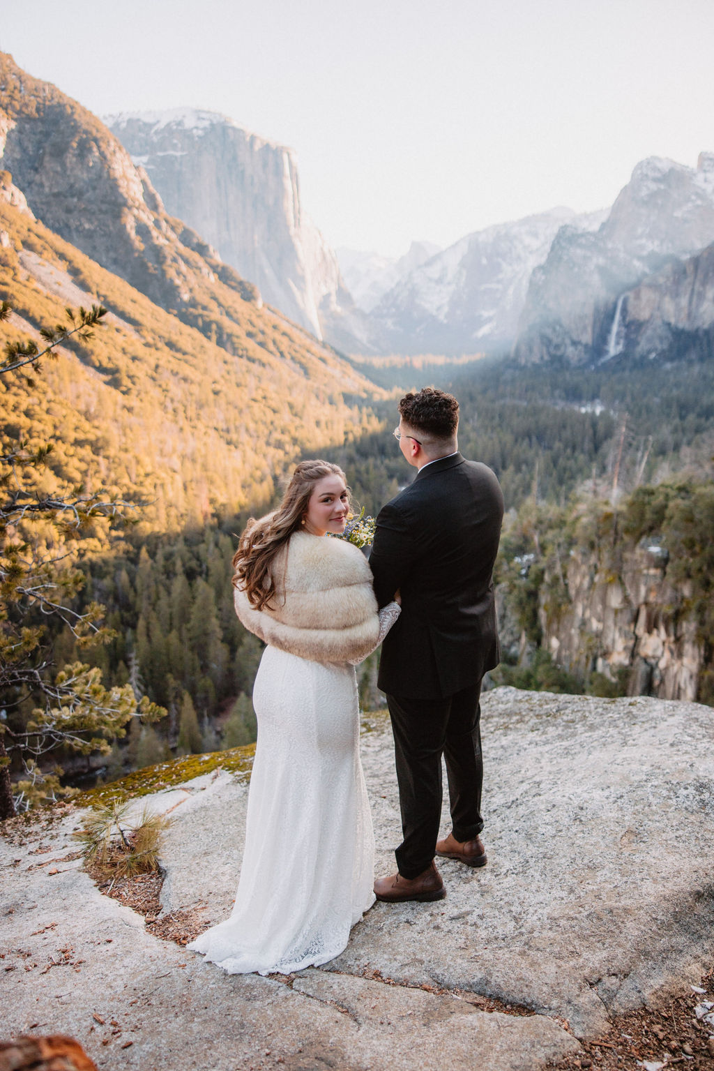 A couple in wedding attire stands smiling on a rock with a mountainous landscape in the background. How to Include Friends & Family in Your Elopement